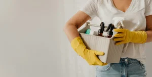 A person holds a box full of cleaning supplies. They are wearing rubber gloves.